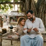 गाँव के आँगन में अपनी छोटी भांजी के साथ बैठा एक स्नेही मामा, उसे कलम बनाना सिखाते हुए, ग्रामीण परिवेश में प्रेम, सुरक्षा और बचपन की यादों से भरा भावनात्मक दृश्य।