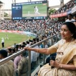 Asha Bhosle क्रिकेट स्टेडियम में बैठकर मैच देखते हुए, पृष्ठभूमि में Sachin Tendulkar बल्लेबाजी करते हुए दिखाई दे रहे हैं.