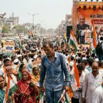 झुकी हुई रीढ़ के प्रतीक के रूप में लोकतंत्र, चुनावी रैली, भीड़ और मंच पर खड़े नेता का व्यंग्यात्मक दृश्य