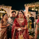 लाल जोड़े में सजी एक भारतीय दुल्हन विदाई के समय आँसुओं से भरी आँखों के साथ पीछे की ओर चावल फेंकती हुई दिख रही है, पास में खड़े माता-पिता भावुक हैं और शादी का सजा हुआ मंडप पृष्ठभूमि में दिखाई दे रहा है।